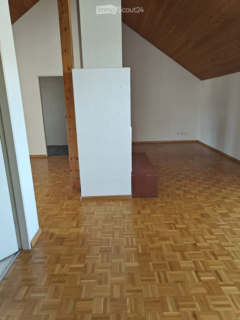 Empty room with wooden herringbone flooring, white walls, and a small red bench against one wall. The room has a slanted wooden ceiling and a doorway leading to another area.