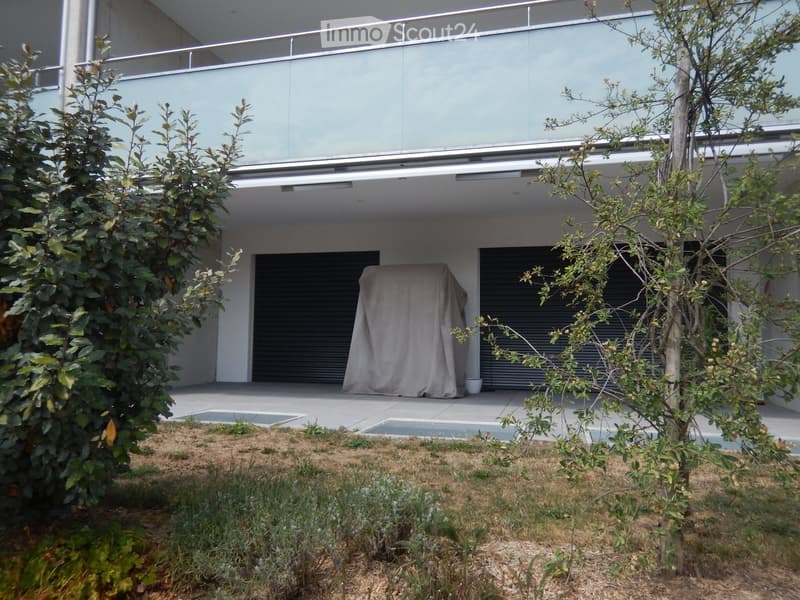 Modern two-story building with a large balcony, covered entrance, and landscaped garden area with small trees and shrubs.