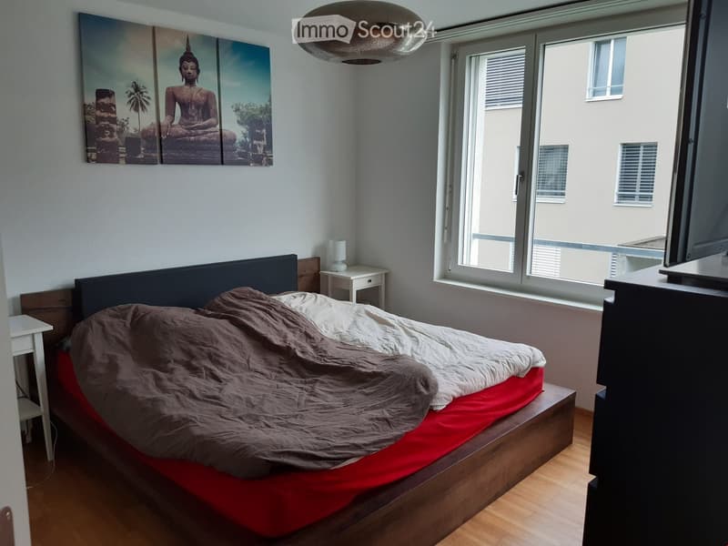 A modern bedroom with a wooden bed, nightstand, lamp, and a large window with blinds. The bed is covered with a brown blanket and a red sheet. A large screen is placed on a stand near the window.