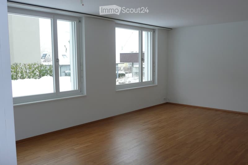 Empty room with wooden flooring and large windows, showing a view of the outside with snow-covered ground and trees.