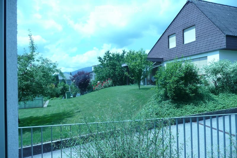 A well-maintained garden with a green lawn, various trees, shrubs, and a metal railing in the foreground. A house with a dark roof and white walls is visible in the background.
