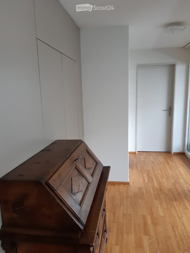 A wooden cabinet with drawers and a slanted top is placed against a white wall in a hallway with wooden flooring and white walls.
