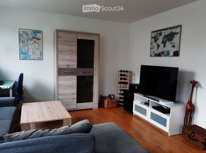 Modern living room with light wood flooring, a grey sofa, a wooden coffee table, a flat-screen TV on a white stand, a wine rack, a tall cabinet, and decorative maps on the walls.