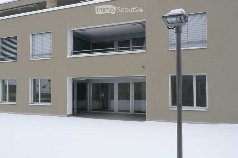 Modern building exterior with large windows, a covered entrance, and a balcony area. Snow covers the ground and roof.