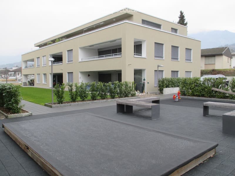 Modern two-story building with beige facade, multiple windows, balconies, and a landscaped courtyard with benches and greenery.