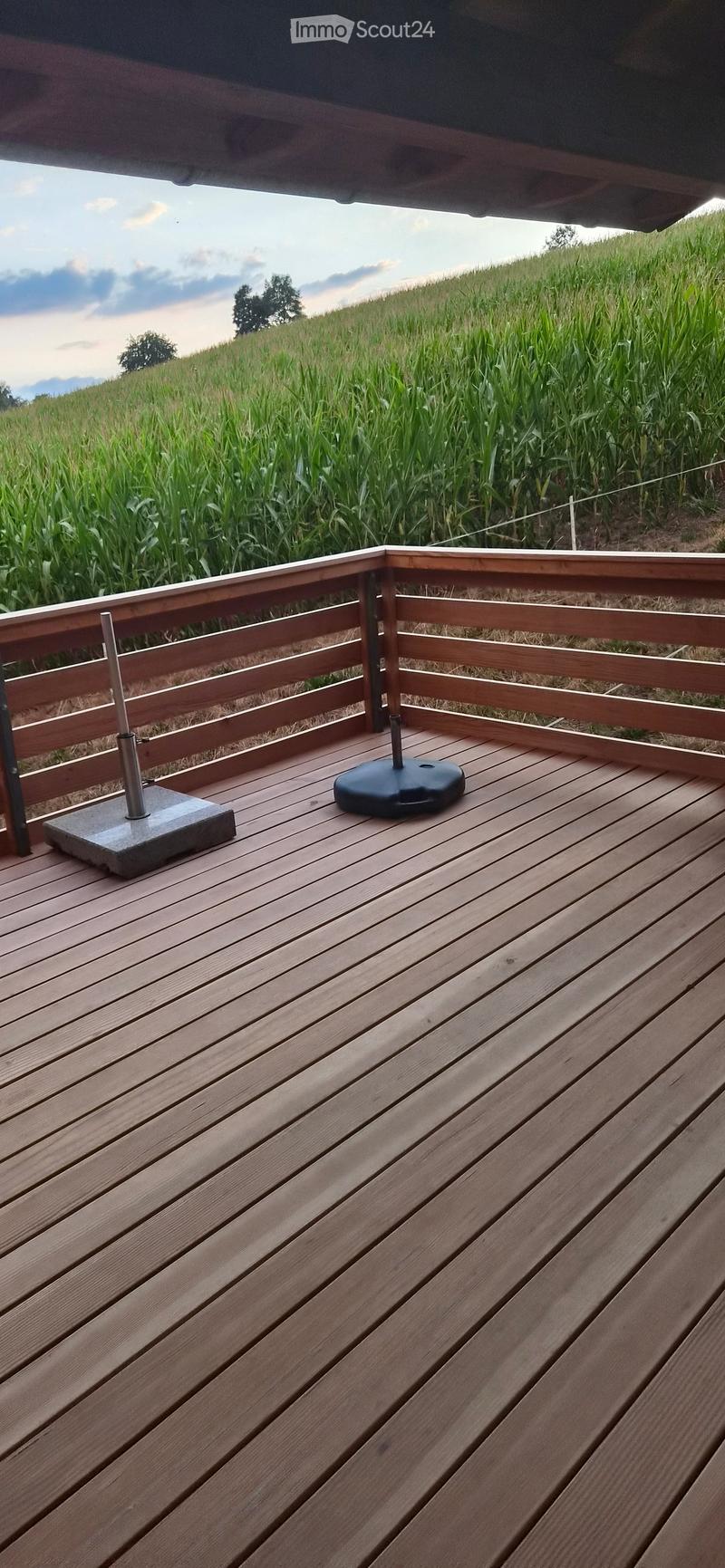 Wooden deck with railing, featuring a concrete base for a parasol and a black parasol base, overlooking a cornfield.