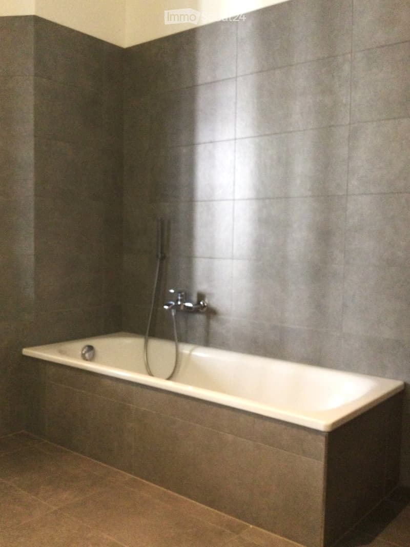 Bathtub with a modern faucet, surrounded by grey tiled walls and a tiled floor.