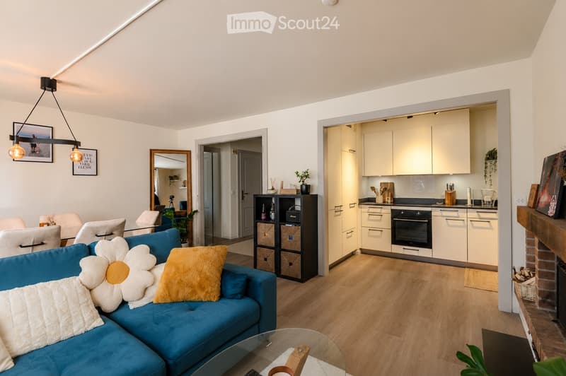 Modern living room with a blue sectional sofa, glass coffee table, shelves, and an open kitchen area with white cabinets and an oven.
