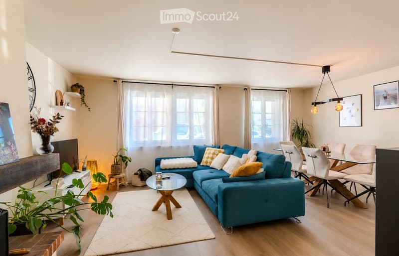 Modern living room with a blue sectional sofa, coffee table, dining area with chairs and table, pendant lighting, and decorative plants.