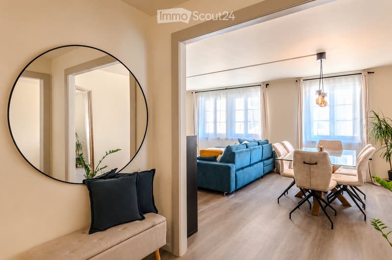 Modern living and dining area with a blue sofa, glass dining table, chairs, a large round mirror, and a small bench with pillows.
