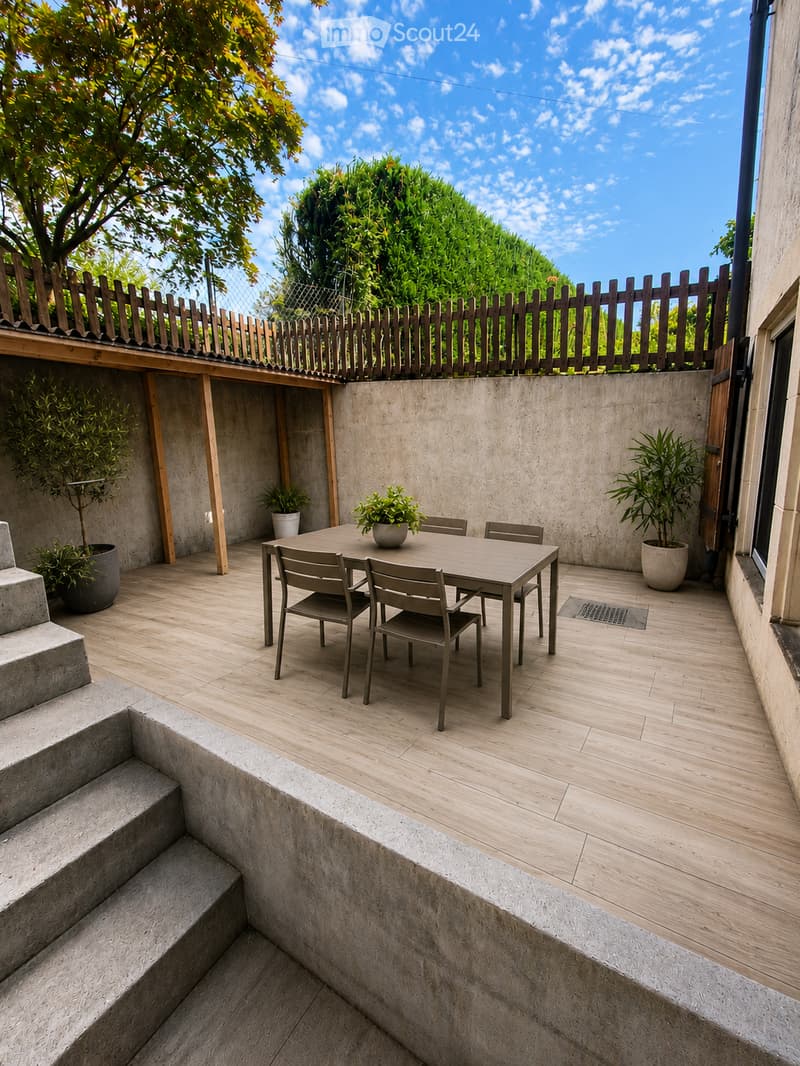 Modern outdoor terrace with wooden flooring, concrete steps, a small table with four chairs, and potted plants. The area is enclosed by a wooden fence and has a clear blue sky view.