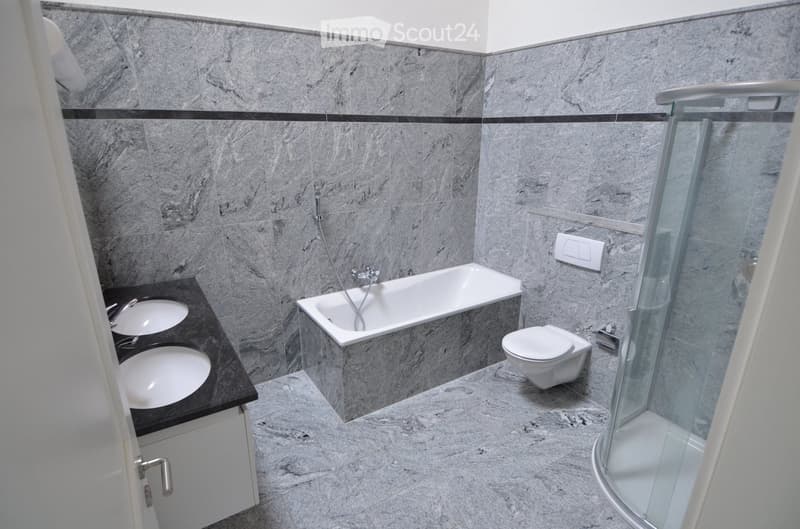 Modern bathroom with marble walls and floors, featuring a bathtub, toilet, double sink, and a glass-enclosed shower.
