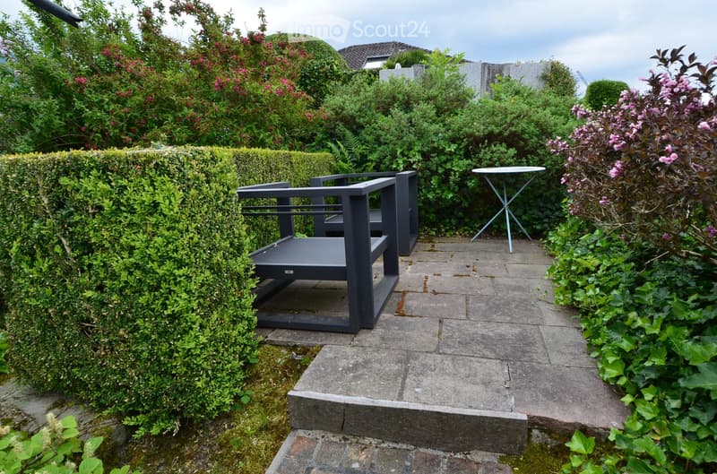 A well-maintained garden with a neatly trimmed hedge, a modern dark grey bench, a small round table, and lush greenery including flowering shrubs and climbing plants.