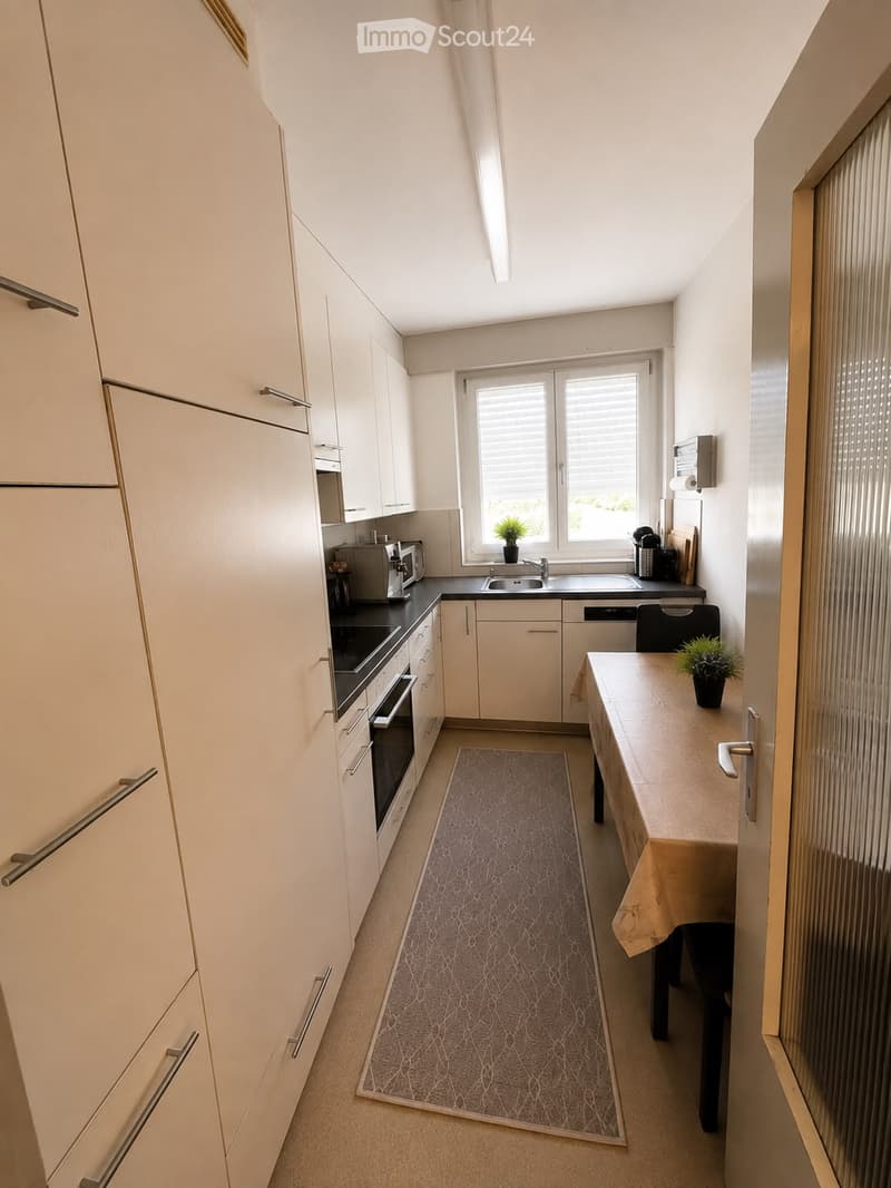 Modern kitchen with light-colored cabinets, stainless steel appliances, a window with blinds, and a small dining area with a table and chair.