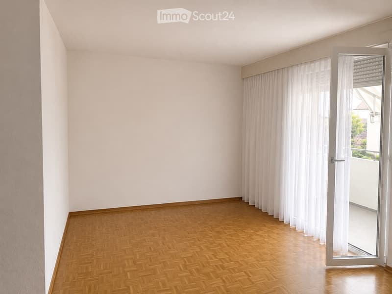 Empty room with wooden herringbone flooring, white walls, and a large window with white curtains leading to a balcony.