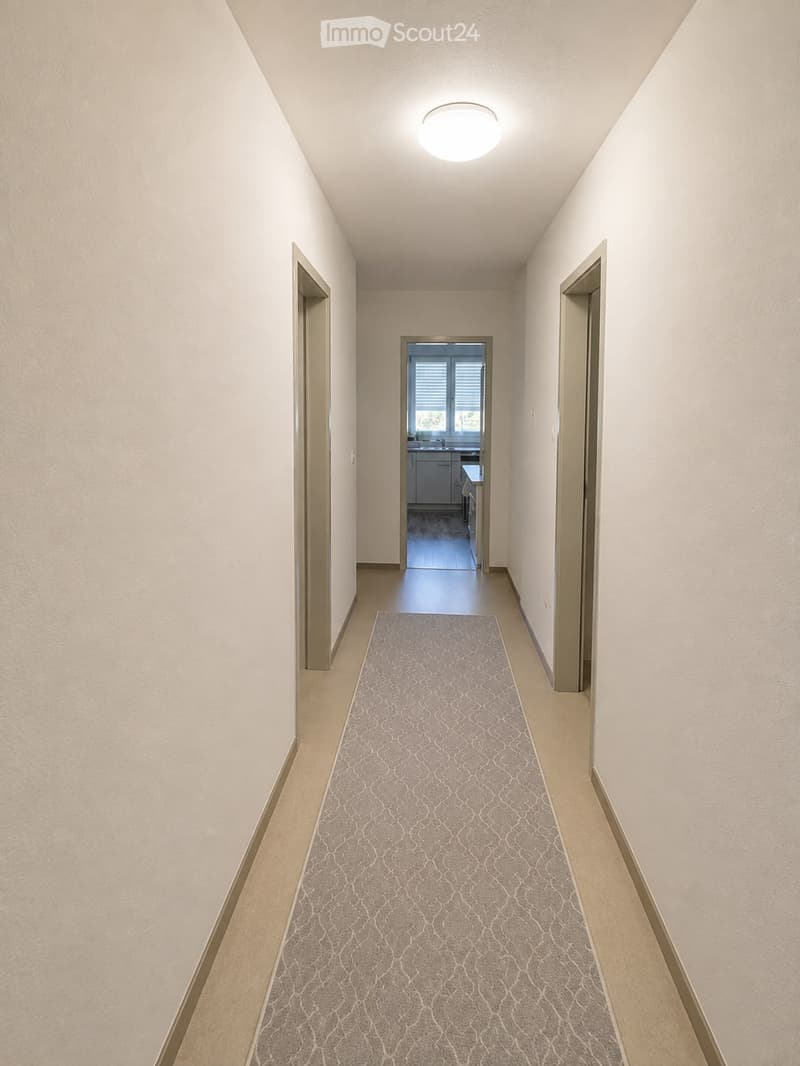 A clean, well-lit hallway with beige walls, a patterned carpet, and a ceiling light fixture. The hallway leads to an open doorway showing a glimpse of a kitchen area with cabinets and a countertop.