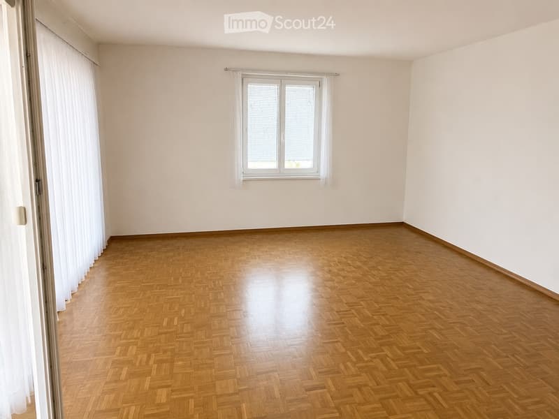 Empty room with wooden herringbone flooring, white walls, and a large window with white blinds and curtains.