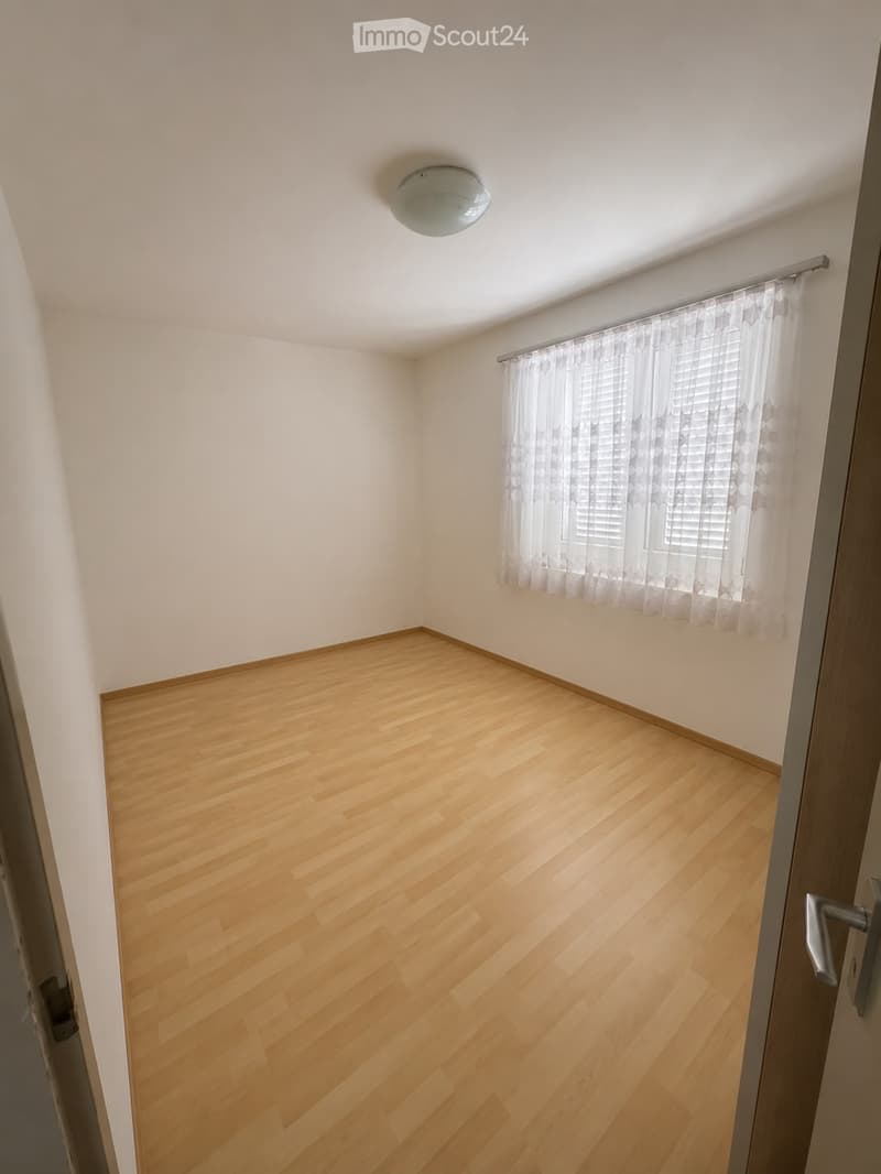 Empty room with light wooden flooring, white walls, a ceiling light fixture, and a window with sheer curtains.