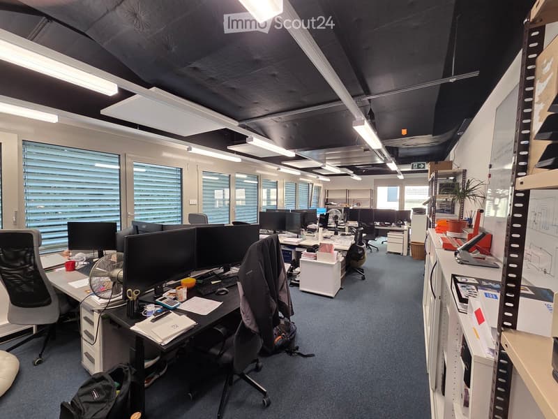 Office space with desks, chairs, multiple computer monitors, keyboards, mice, fans, cups, backpacks, and a plant. Large windows with blinds provide natural light.