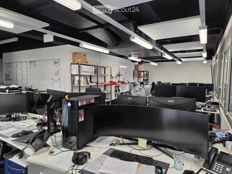 Office space with multiple desks, computers, chairs, and office supplies. Shelves hold books and files, and a whiteboard is visible on the wall.