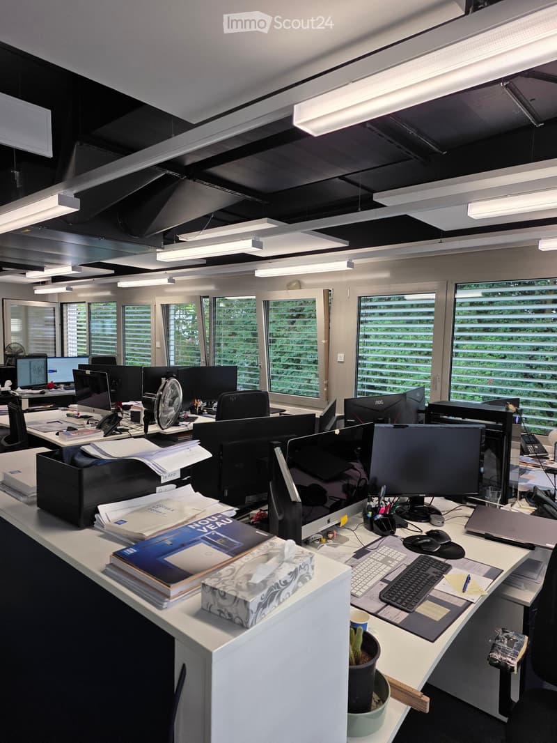 Office space with multiple desks, computers, chairs, and office supplies. Large windows provide natural light, and the room is equipped with modern office furniture and equipment.