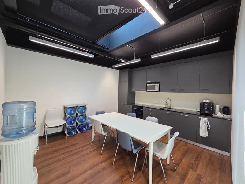 Modern kitchen area with grey cabinets, white countertop, sink, microwave, water dispenser, and a white table with chairs.