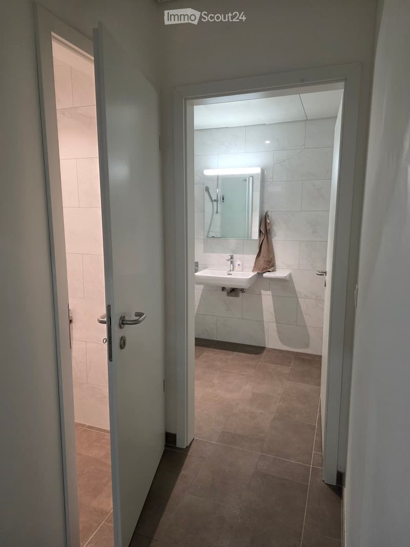 Modern bathroom with a white sink, large mirror, and a towel hanging on the side. The floor is tiled in a warm brown color, and the walls are covered with white tiles.