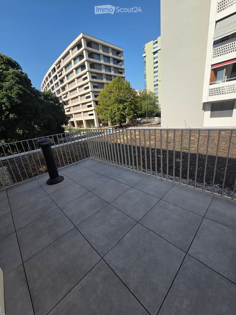 Balcony with tiled floor, metal railing, and a black lamp post, overlooking a landscaped area with trees and buildings.