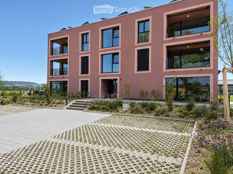 Modern two-story building with pink facade, large windows, and balconies. The entrance features a small garden with plants and flowers, and a paved walkway leads to the building.