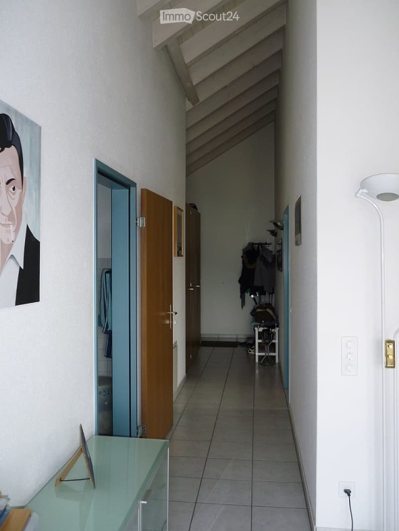 A narrow hallway with tiled flooring, white walls, and a slanted ceiling. Features include a glass console table with a small decorative item, a painting on the wall, and several doors leading to other rooms.