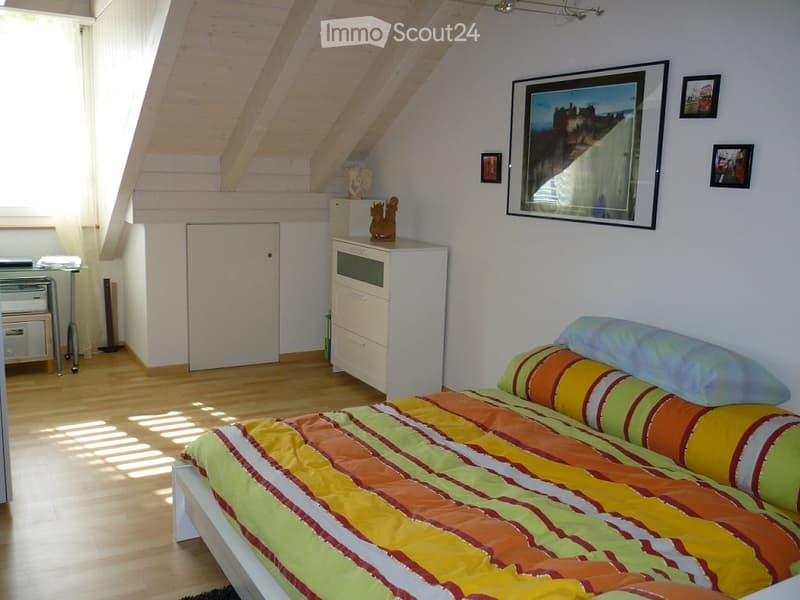 Bright bedroom with wooden floors, slanted ceiling beams, and a colorful striped bedspread. Includes a nightstand, mirror, and decorative items on a cabinet.