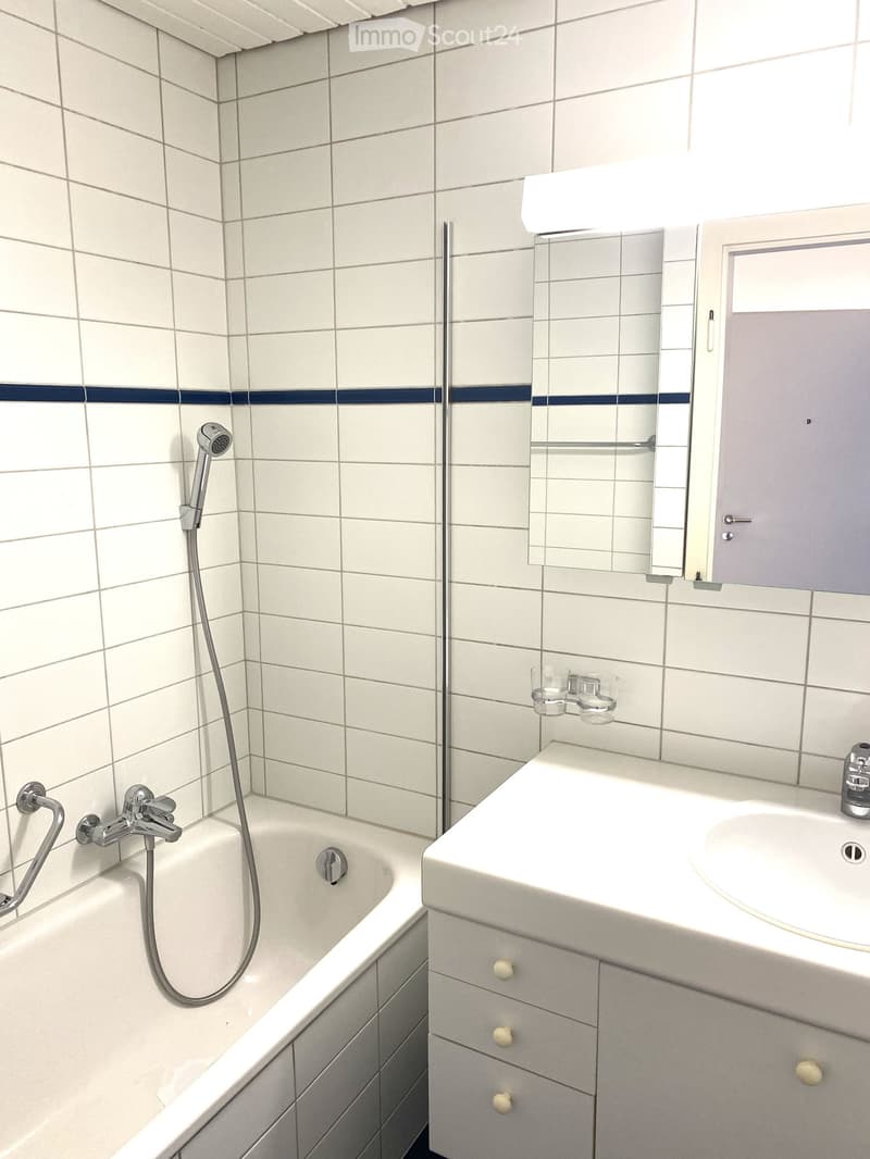 White bathroom with white tiles and blue border, bathtub with showerhead, mirror, sink, and drawers.