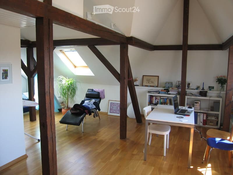 Bright, wooden-floored room with slanted ceiling beams, skylight window, laptop on desk, chair, shelf with books, plant, and decorative items.