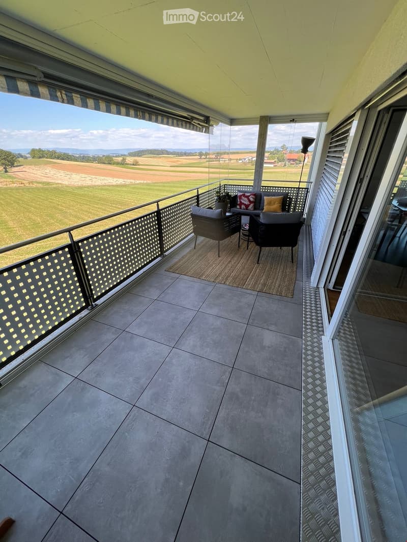 Balcony with tiled floor, glass railing, sofa, chairs, small table, and potted plant, overlooking a scenic countryside view.