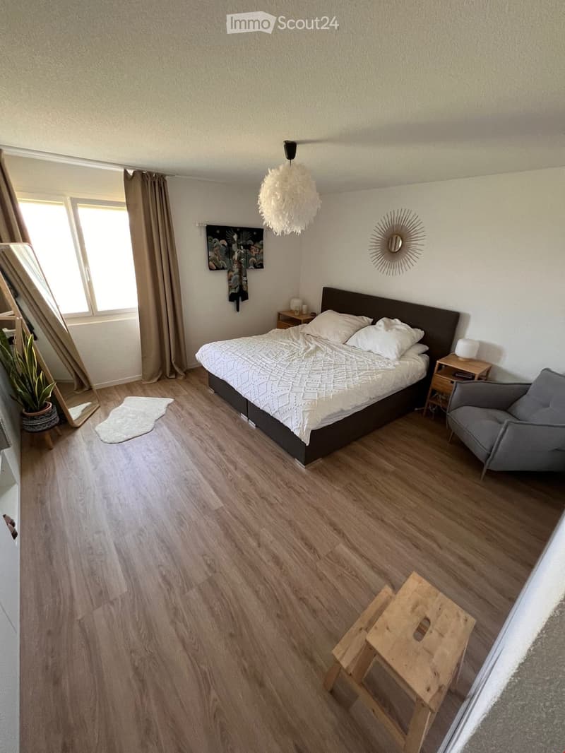 Modern bedroom with wooden flooring, a large bed with white bedding, a nightstand with a lamp, a grey armchair, a small wooden stool, a mirror, a decorative lamp, and a potted plant near the window.