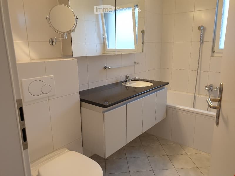 Modern bathroom with white tiles, a black countertop sink, a bathtub, a toilet, and a mirrored cabinet with a round mirror.