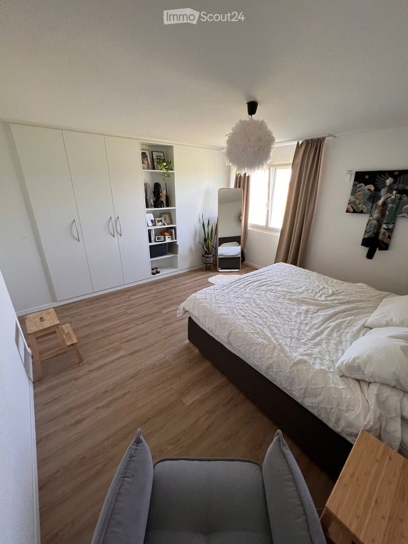 Modern bedroom with a large bed, white bedding, grey armchair, wooden floor, white wardrobe, mirror, and decorative items.
