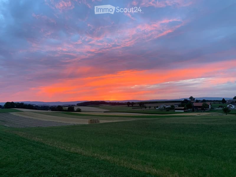 Rural landscape with green fields, scattered houses, and a colorful sunset sky.