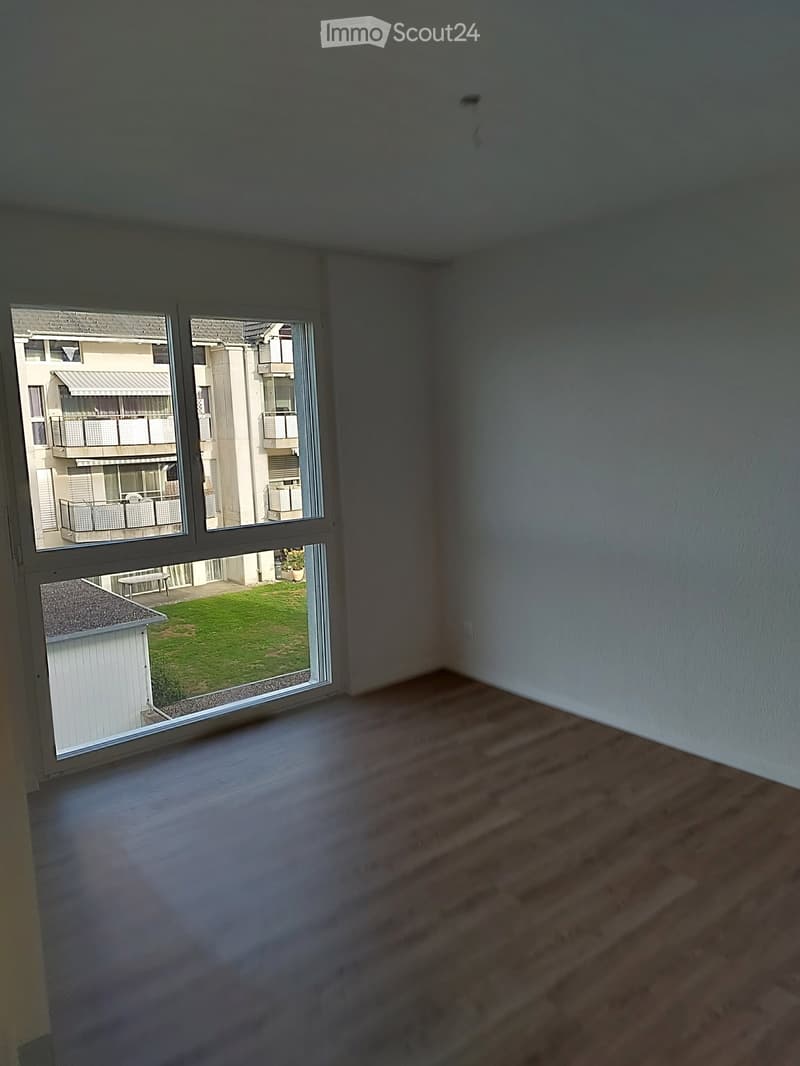 Empty room with wooden flooring and large windows offering views of a green lawn and neighboring buildings.