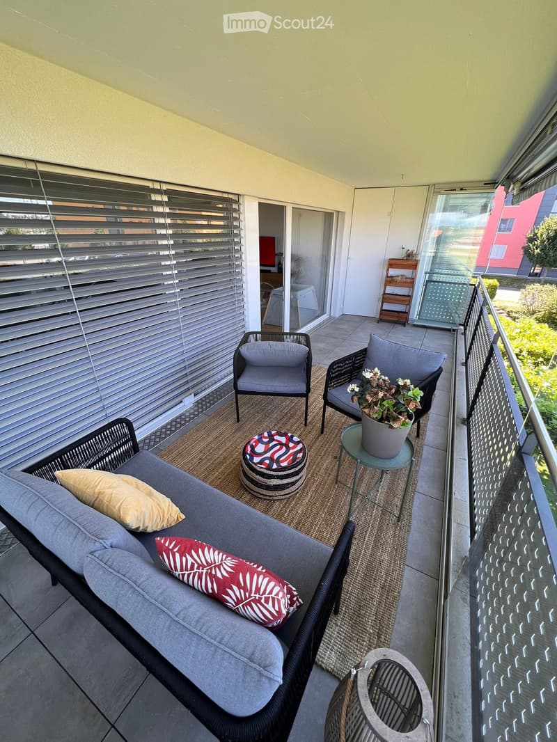 Balcony with a grey wicker sofa, two chairs, a small table with a potted plant, a round stool, and a woven rug.
