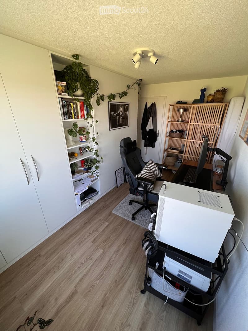 A well-organized home office with a wooden floor, a large bookshelf, a desk with a computer, a comfortable chair, and various decorative items including plants and artwork.