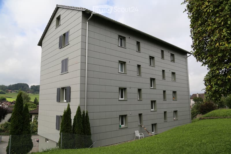 Modern multi-story building with grey siding, multiple windows, and a raised ground floor entrance. The building is surrounded by greenery and a fence.