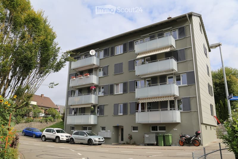 Four-story apartment building with multiple balconies, satellite dishes, and several parked cars in front.