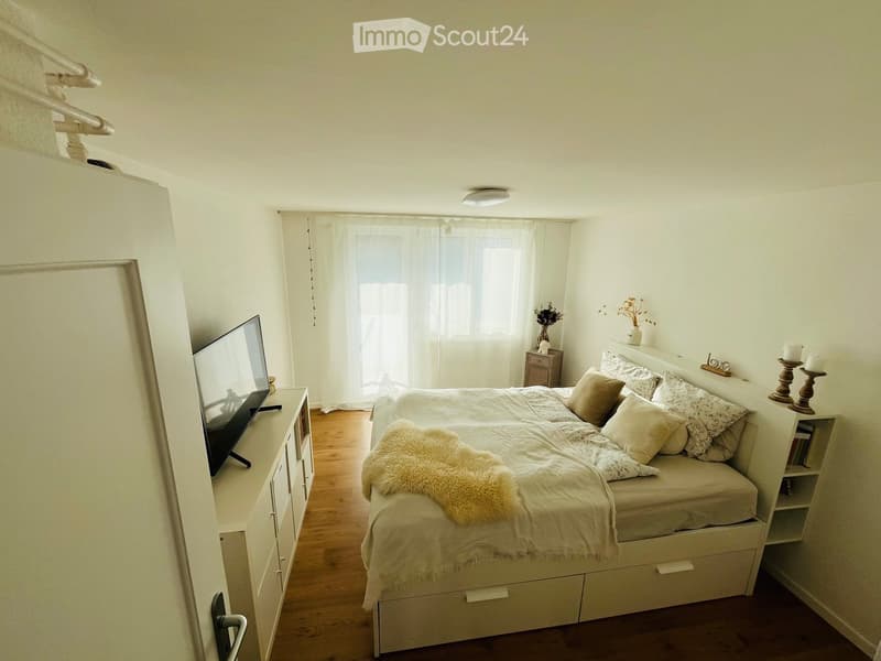 Cozy bedroom with a large bed, nightstand, television, and decorative items. The room features wooden flooring and large windows with white curtains.
