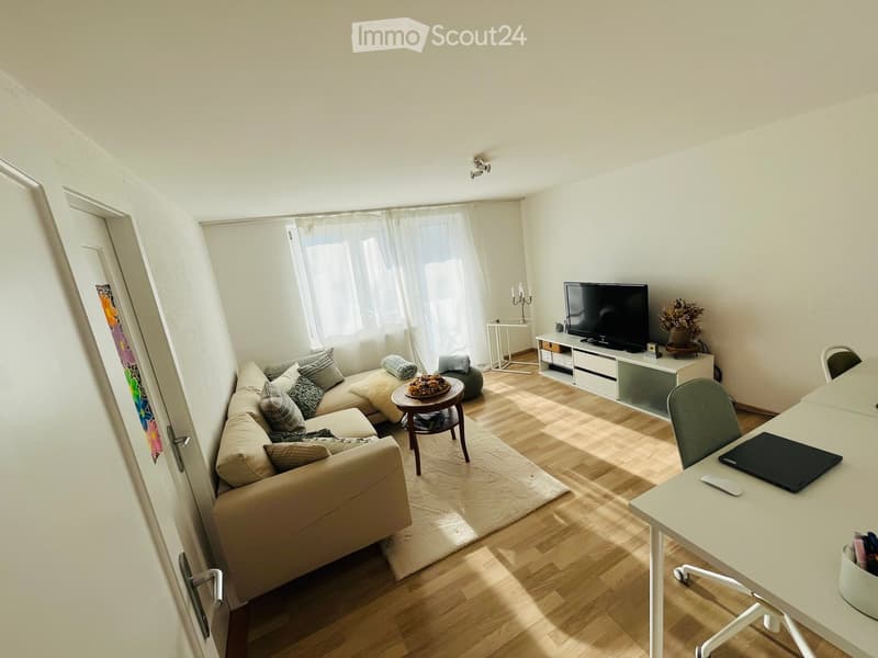 A modern living room with a beige sofa, coffee table, TV stand, and a small desk with a laptop. The room features wooden flooring, large windows with white curtains, and a small side table with a candle holder and plant.