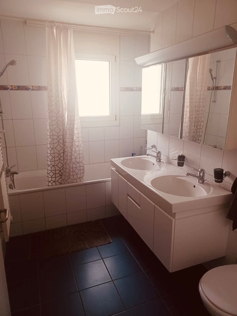 Modern bathroom with white tiles, a bathtub, double sink, mirror, and a toilet. Features a showerhead and a patterned shower curtain.