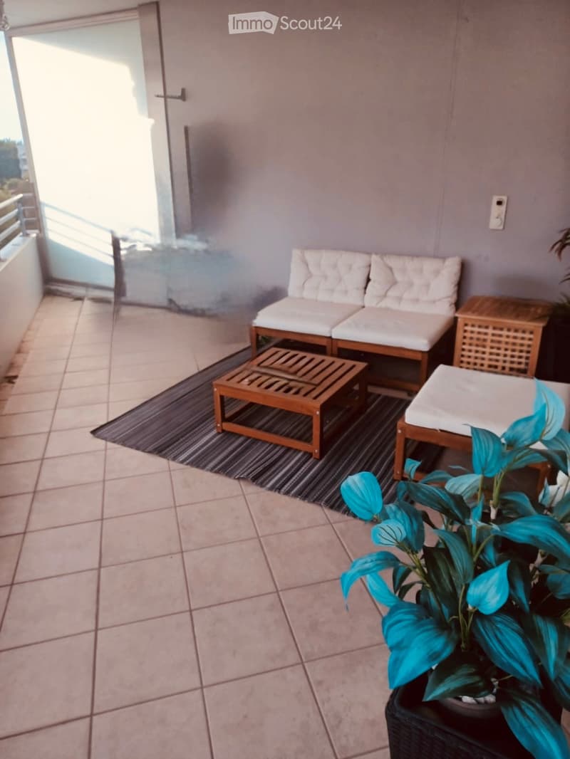 Balcony with tiled floor, white cushioned sofa, wooden coffee table, small wooden side table, and potted plant.