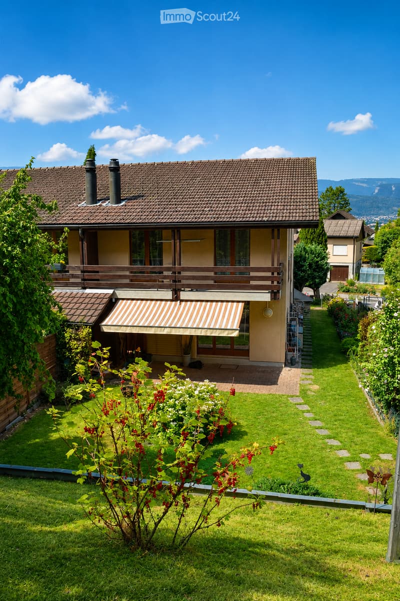 Two-story house with a tiled roof, multiple chimneys, and a balcony. The house is surrounded by a well-maintained garden with a lawn, flower beds, and a pathway leading to the entrance.