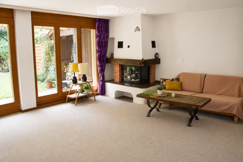 Bright living room with a beige sofa, coffee table, fireplace, side table with lamps, and plants. Large sliding glass doors lead to a garden.