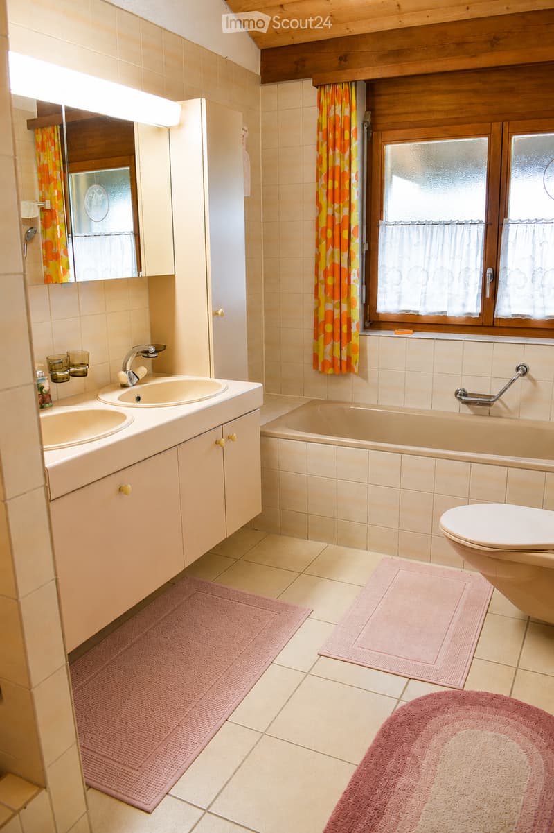 Bathroom with beige tiles, double sink, bathtub, toilet, mirror, and pink rugs.